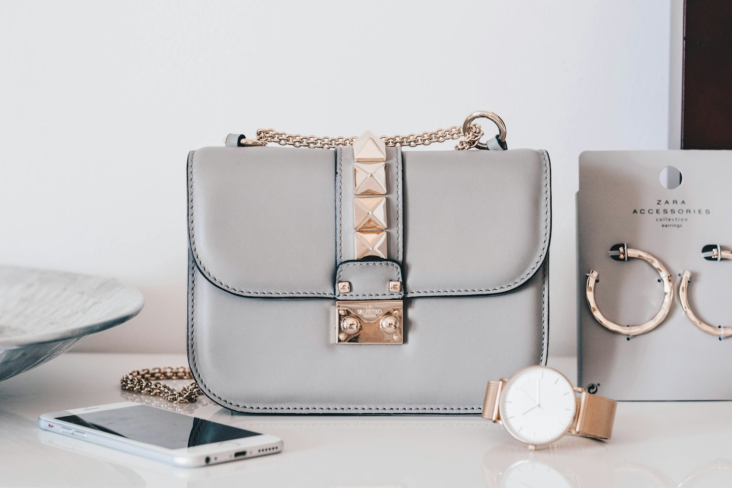Borsa grigia, smartphone, orologio e orecchini a cerchio su una superficie bianca.