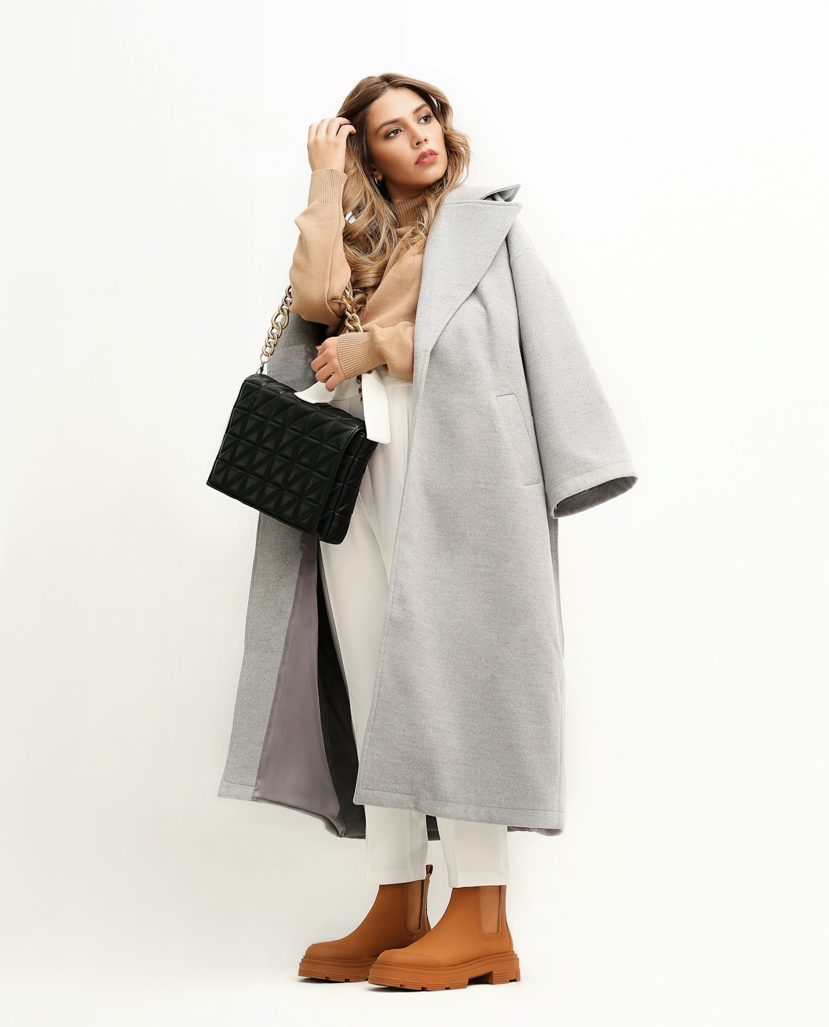 Donna con cappotto grigio e pantaloni bianchi tiene una borsa nera.